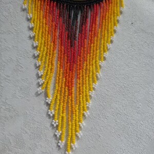 May include: A beaded necklace with a circular top featuring a design in red, yellow, and black. The necklace has long, beaded fringe in shades of yellow, orange, and red, with white floral accents at the ends. The necklace is likely handmade.