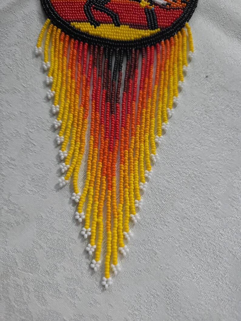May include: A beaded necklace with a circular top featuring a design in red, yellow, and black. The necklace has long, beaded fringe in shades of yellow, orange, and red, with white floral accents at the ends. The necklace is likely handmade.