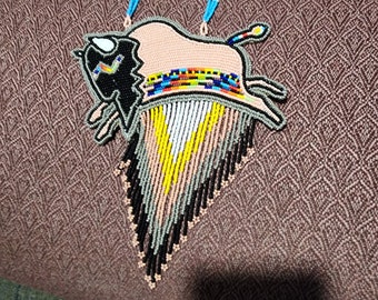 May include: A beaded necklace featuring a bison design. The bison is rendered in black, tan, and colorful beads, with a fringe of beads in various colors. The necklace has a beaded chain with blue and white accents.