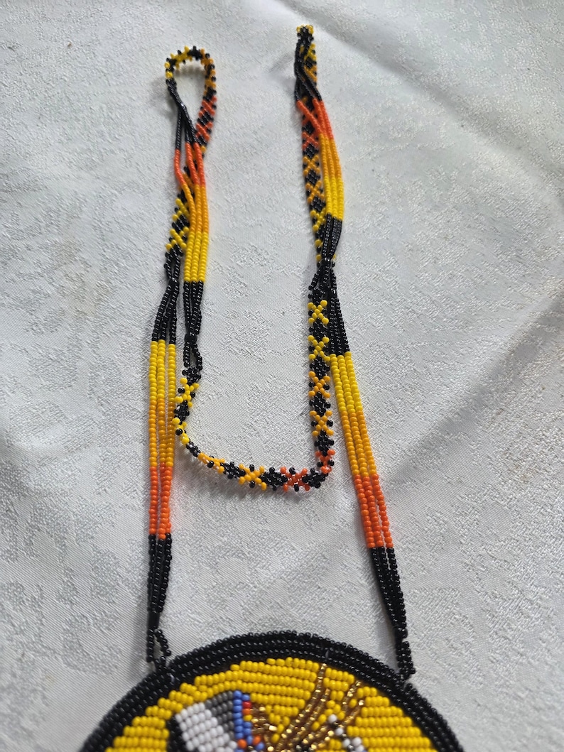 May include: A beaded necklace with a circular pendant. The necklace features strands of yellow, orange, and black beads. The pendant has a yellow background with a black border and colorful details. The necklace is likely handmade.