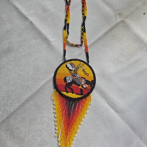 May include: Handmade beaded necklace featuring a circular pendant with a Native American design. The pendant is yellow with a black border and a beaded image of a horse and rider. The necklace has a beaded fringe in yellow, orange, and red.
