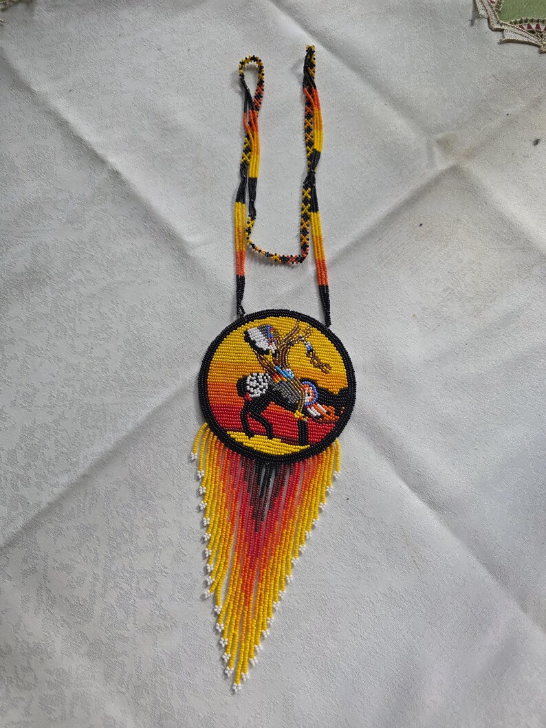 May include: Handmade beaded necklace featuring a circular pendant with a Native American design. The pendant is yellow with a black border and a beaded image of a horse and rider. The necklace has a beaded fringe in yellow, orange, and red.