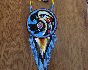 May include: A beaded necklace with a circular pendant featuring an eagle design. The pendant is primarily blue, black, white, red, and yellow. The necklace has a blue beaded fringe and a blue, red, and yellow beaded chain.