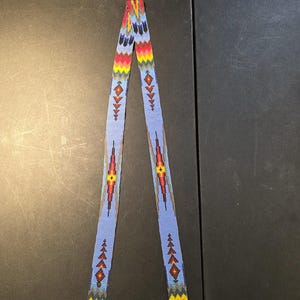 May include: A beaded strap with a light blue background and colorful geometric patterns. The strap features red, yellow, and blue designs, with diamond and triangle shapes. The ends are finished with yellow and red beads.