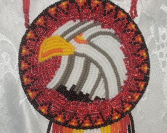 May include: A beaded necklace with a round pendant featuring an eagle design. The pendant is made with red, white, orange, yellow, and black beads. The necklace is made with red, white, and yellow beads.