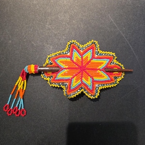 May include: A handcrafted beaded hair barrette in a starburst design. The barrette features a vibrant color palette of red, orange, yellow, and blue beads. A wooden stick is used to secure the barrette in the hair. The barrette has a beaded tassel.