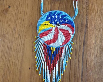 May include: A beaded necklace with a round pendant featuring an eagle design with red, white, and blue colors. The pendant has a fringe of beads in the same colors.