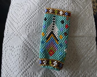 May include: A rectangular lighter case covered in intricate beadwork featuring a turquoise background. The design includes a central white triangular motif with geometric patterns in red, yellow, blue, and purple, bordered by metallic gold bead bands.