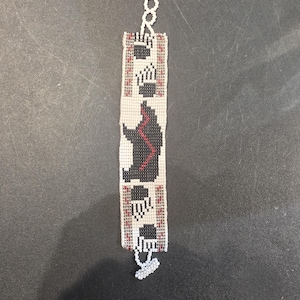 May include: Handmade beaded bracelet featuring a central bear design in black and red. The bracelet has a white background with black paw prints and red accents. The bracelet is approximately 7 inches long.