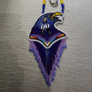 May include: A beaded necklace featuring an eagle head design. The necklace is primarily blue, purple, and white, with yellow accents. The eagle's head is detailed with intricate beadwork, and the pendant has a fringe of beads at the bottom.
