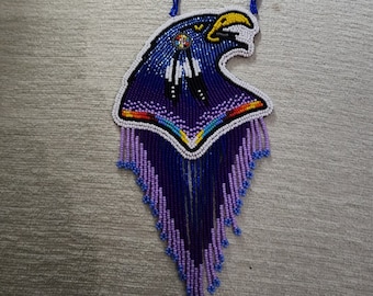 May include: A beaded necklace featuring an eagle head design. The necklace is primarily blue, purple, and white, with yellow accents. The eagle's head is detailed with intricate beadwork, and the pendant has a fringe of beads at the bottom.