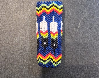 May include: A rectangular beaded item with a blue background and a zig-zag pattern in yellow, red, and green. Two white and yellow feather designs are centered on the blue background. The item is likely a bracelet or a similar accessory.
