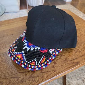May include: Black baseball cap with a beaded brim. The brim features a geometric pattern in red, blue, yellow, and white beads against a black background. The cap is sitting on a wooden table.