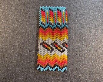 May include: Rectangular beaded item with a vibrant pattern. The design features rows of small beads in shades of blue, red, yellow, orange, white, and black. The pattern includes zigzag and geometric shapes, creating a colorful and textured appearance.