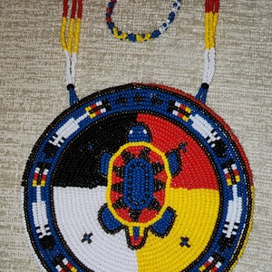May include: A beaded necklace with a round pendant featuring a turtle design in the center. The pendant is surrounded by four colored sections: black, white, yellow, and red. The necklace is made of blue, white, red, and yellow beads.