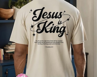 May include: Cream-colored t-shirt with the text "Jesus is King" in a stylized black font, encircled by a curved line and stars. Below the text is a Bible verse from 1 Timothy 6:15.