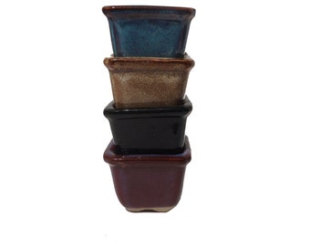 May include: Four square ceramic pots stacked on top of each other. The top pot is blue, the second pot is brown, the third pot is black, and the bottom pot is burgundy.