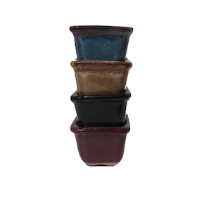May include: Four square ceramic pots stacked on top of each other. The top pot is blue, the second pot is brown, the third pot is black, and the bottom pot is burgundy.