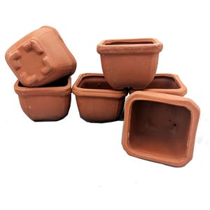 May include: A collection of small, square terracotta pots. The pots are stacked and arranged, showcasing their simple, yet functional design. The unglazed clay has a warm, earthy tone, perfect for small plants or succulents.