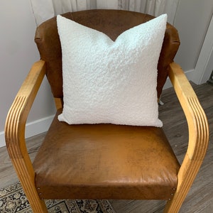 May include: A white fluffy pillow sits on a brown leather armchair with wooden arms. The chair is in a room with a light wood floor.