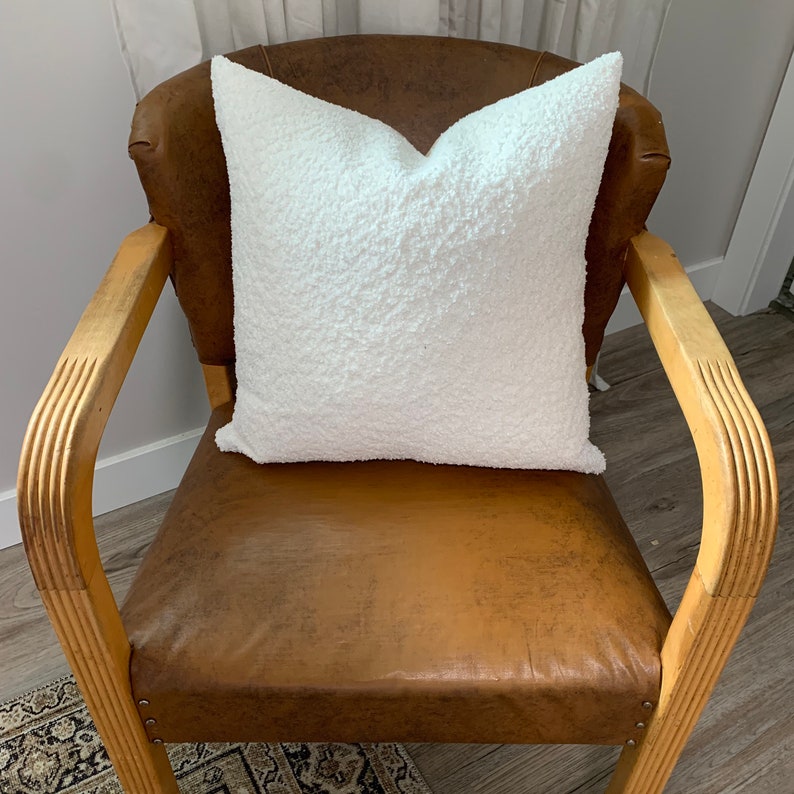 May include: A white fluffy pillow sits on a brown leather armchair with wooden arms. The chair is in a room with a light wood floor.