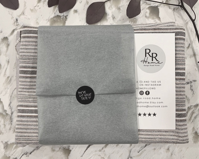 May include: A gray and white striped throw blanket is folded and wrapped in gray tissue paper. The tissue paper has a black circle sticker with the text "You've Got Great Taste" on it. The blanket is packaged with a white card with the text "RR Home" and social media information.