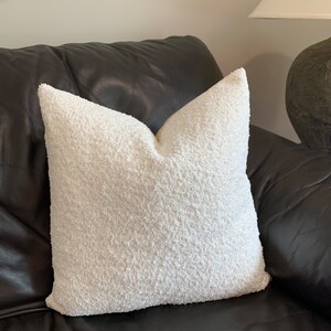 May include: A white fluffy pillow with a soft, textured surface. The pillow is placed on a black leather couch.