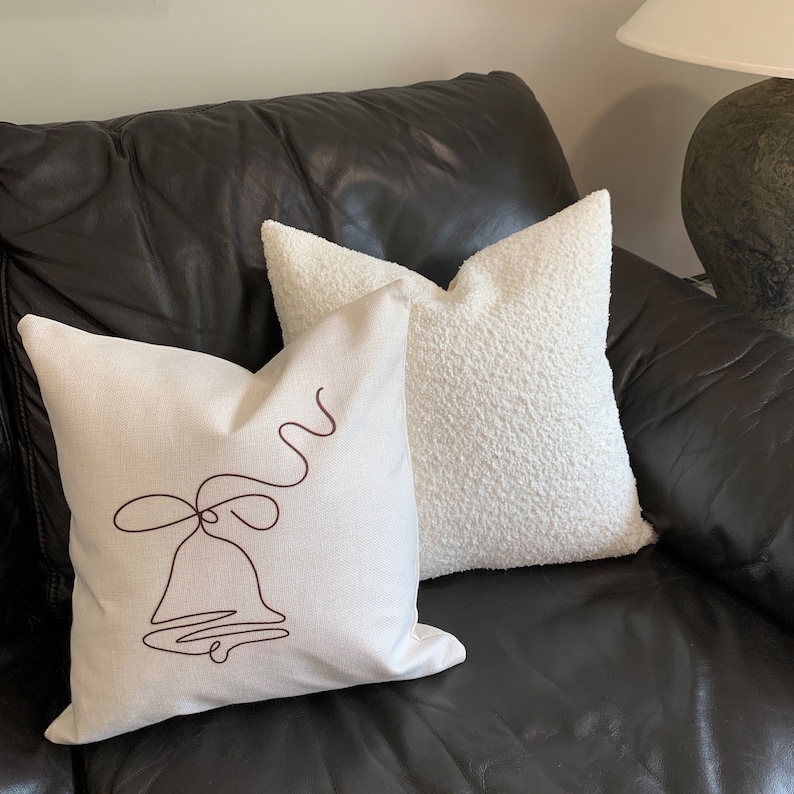 May include: A white throw pillow with a brown bell design on a white linen pillow. Both pillows are on a black leather couch.