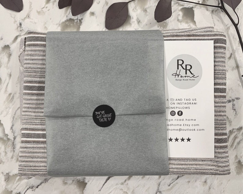 May include: A gray and white striped throw blanket is folded and wrapped in gray tissue paper. The tissue paper has a black circle sticker with the text "You've Got Great Taste" on it. The blanket is packaged with a white card with the text "RR Home" and social media information.