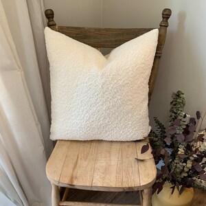 May include: A white fluffy pillow sits on a wooden chair with a light brown finish. The chair has a carved wooden top and a simple design.
