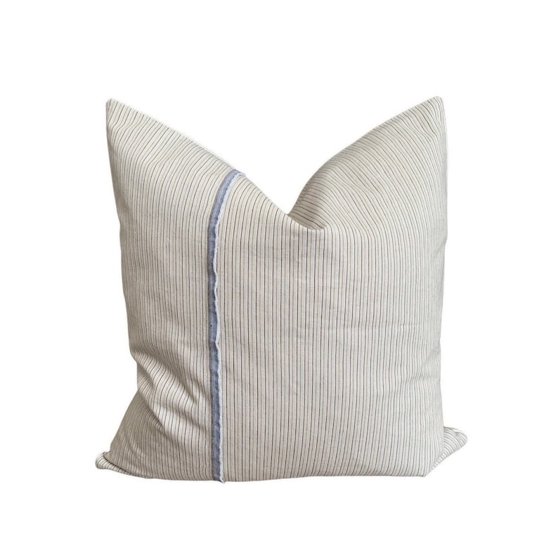 May include: A cream-colored pillow with thin black stripes and a blue and white trim down the center.
