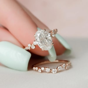 Può includere: Un anello di fidanzamento in oro rosa con un grande diamante di forma ovale e una sottile fascia di diamanti. L'anello è incastonato con diamanti più piccoli sulla fascia.