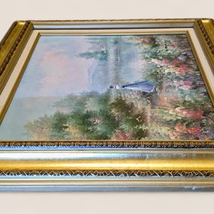 May include: A framed landscape painting featuring a figure in a blue dress walking near a body of water. The artwork is surrounded by a gold-colored ornate frame with a white inner border. The painting depicts a serene outdoor scene.
