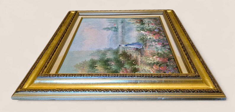 May include: A framed landscape painting featuring a figure in a blue dress walking near a body of water. The artwork is surrounded by a gold-colored ornate frame with a white inner border. The painting depicts a serene outdoor scene.