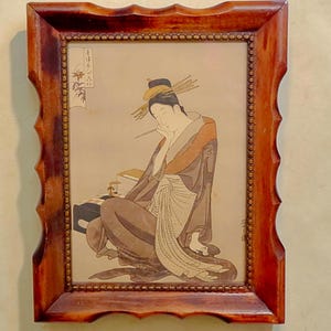Könnte beinhalten: Ein gerahmter Druck mit einer Illustration im Ukiyo-e-Stil, die eine sitzende Figur mit aufwendiger Frisur zeigt, die einen dünnen Pinsel hält. Die Figur trägt ein dunkles Gewand mit einer hellen Schärpe. Der Rahmen besteht aus poliertem braunem Holz mit gewelltem Rand.