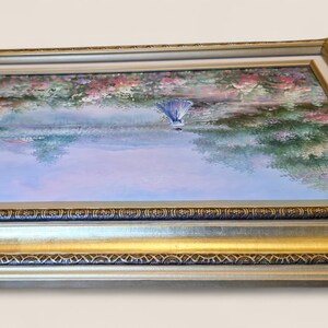 May include: A framed landscape painting featuring a serene scene with a figure in a blue dress. The artwork is set within a gold and silver ornate frame. The painting depicts a waterside scene with trees and flowers.