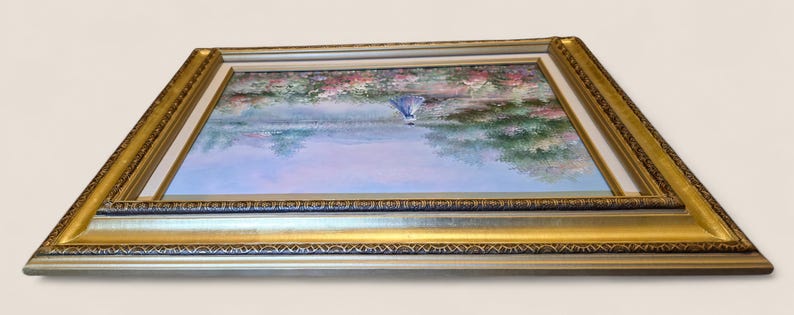 May include: A framed landscape painting featuring a serene scene with a figure in a blue dress. The artwork is set within a gold and silver ornate frame. The painting depicts a waterside scene with trees and flowers.