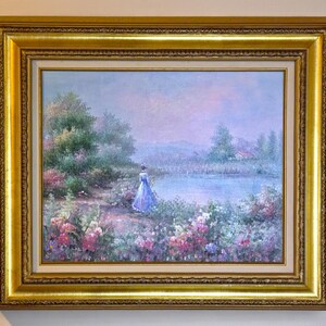 May include: Oil painting in a gold frame. The artwork depicts a woman in a blue dress standing near a lake, surrounded by flowers and trees. The painting has a soft, pastel color palette, with a serene and romantic atmosphere.