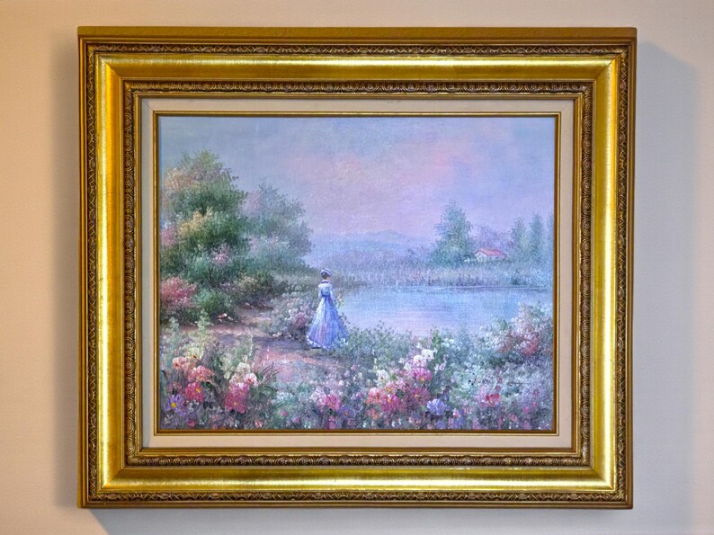 May include: Oil painting in a gold frame. The artwork depicts a woman in a blue dress standing near a lake, surrounded by flowers and trees. The painting has a soft, pastel color palette, with a serene and romantic atmosphere.