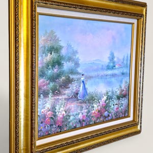 May include: A framed landscape painting featuring a woman in a blue dress by a lake. The artwork has a gold frame with ornate detailing. The scene includes colorful flowers, trees, and a small house.