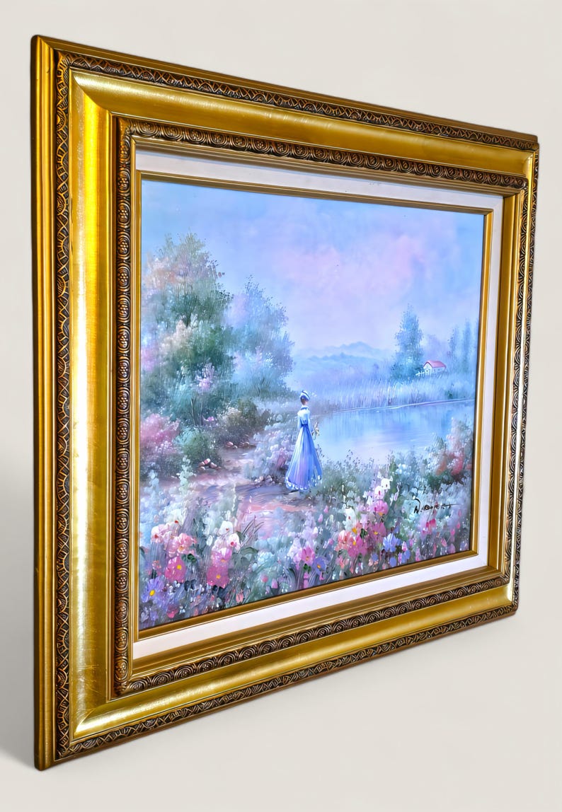 May include: A framed landscape painting featuring a woman in a blue dress by a lake. The artwork has a gold frame with ornate detailing. The scene includes colorful flowers, trees, and a small house.