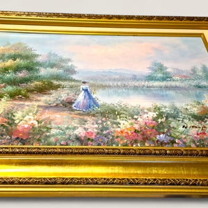 May include: A framed landscape painting featuring a woman in a blue dress near a lake. The artwork is set in a gold ornate frame. The painting depicts a serene outdoor scene with colorful flowers and trees.