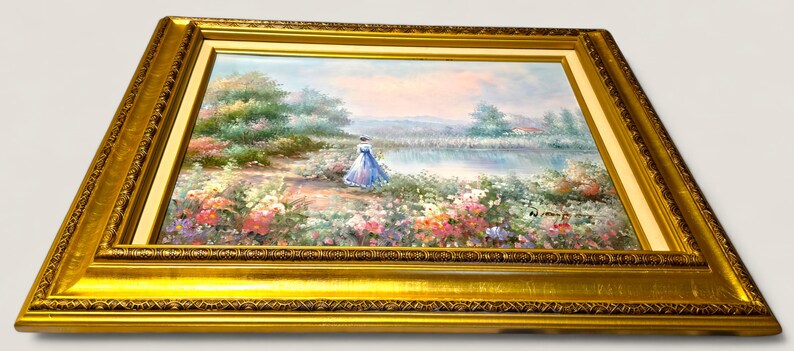 May include: A framed landscape painting featuring a woman in a blue dress near a lake. The artwork is set in a gold ornate frame. The painting depicts a serene outdoor scene with colorful flowers and trees.