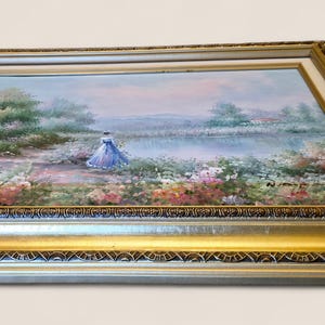 May include: A framed landscape painting featuring a figure in a blue dress near a body of water, surrounded by colorful flowers and greenery. The painting is set within a gold-colored ornate frame, adding a touch of elegance to the artwork.