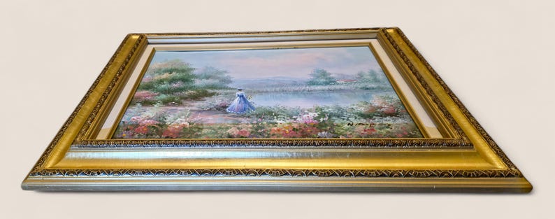May include: A framed landscape painting featuring a figure in a blue dress near a body of water, surrounded by colorful flowers and greenery. The painting is set within a gold-colored ornate frame, adding a touch of elegance to the artwork.