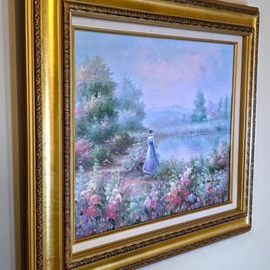 May include: A framed landscape painting featuring a woman in a blue dress standing near a lake. The artwork has a gold ornate frame and depicts a serene scene with colorful flowers, trees, and a small house in the distance. The painting is in a soft, pastel color palette.