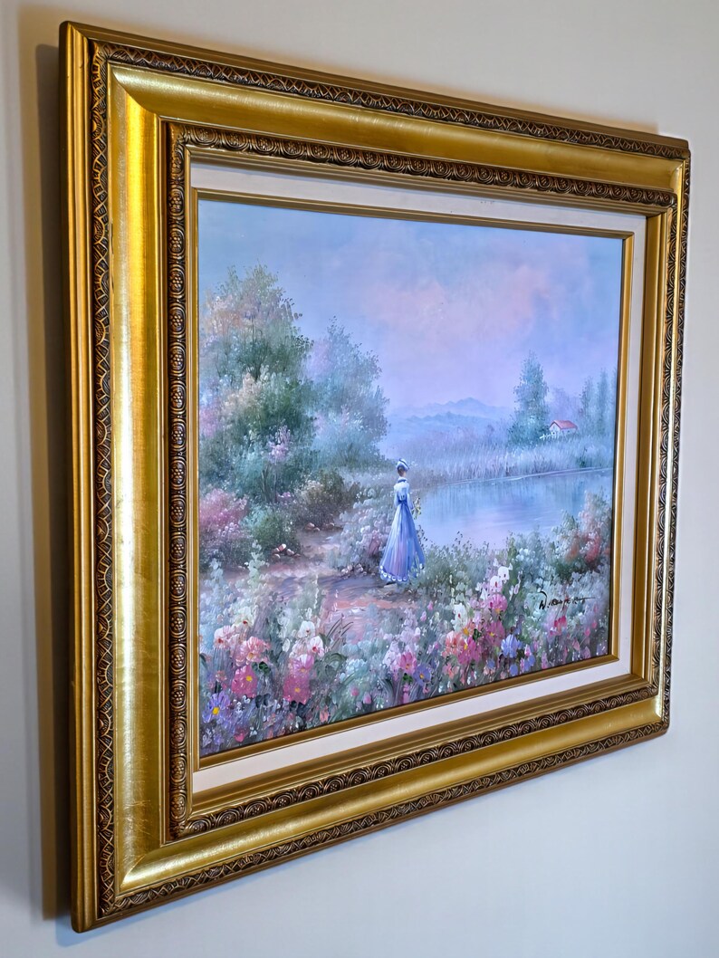 May include: A framed landscape painting featuring a woman in a blue dress standing near a lake. The artwork has a gold ornate frame and depicts a serene scene with colorful flowers, trees, and a small house in the distance. The painting is in a soft, pastel color palette.