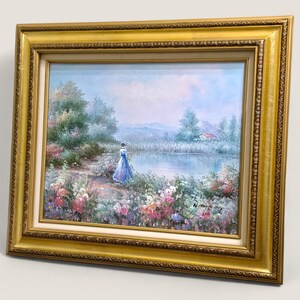 May include: An oil painting in a gold frame depicts a serene landscape with a figure in a blue dress near a lake. The artwork features a woman, flowers, trees, and a house, all under a soft, blue sky.
