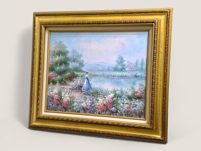 May include: An oil painting in a gold frame depicts a serene landscape with a figure in a blue dress near a lake. The artwork features a woman, flowers, trees, and a house, all under a soft, blue sky.