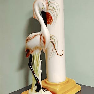 May include: A ceramic vase featuring a white cylindrical column attached to a yellow rectangular base. A sculpted white crane with brown and black accents stands on the base, its neck curved toward the column, which is decorated with painted foliage.